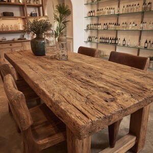 Peut inclure: Une grande table &agrave; manger en bois rustique avec une finition naturelle et vieillie. La table est entour&eacute;e de chaises en bois et de chaises rembourr&eacute;es en cuir. Des bouteilles en verre d&eacute;coratives et un vase avec des fleurs s&eacute;ch&eacute;es sont pos&eacute;s sur la table.