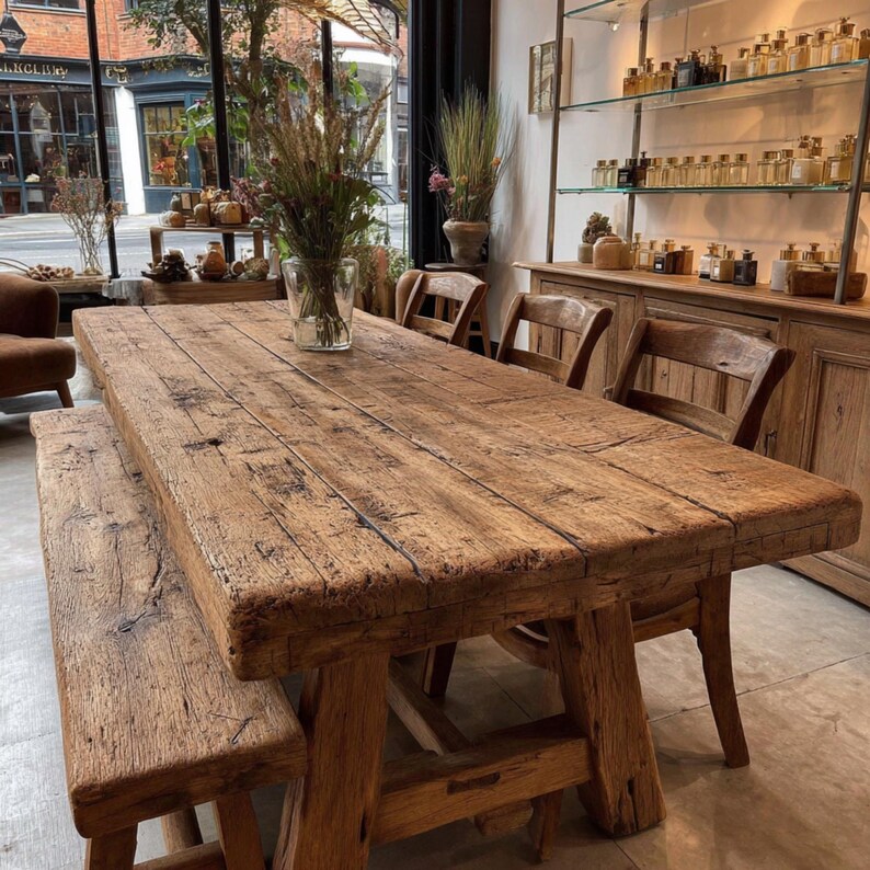 Peut inclure: Une grande table &agrave; manger en bois rustique avec un banc assorti et plusieurs chaises en bois. La table et le banc ont un aspect vieilli et patin&eacute;, mettant en valeur le grain naturel du bois. La pi&egrave;ce comprend &eacute;galement un vase en verre avec des fleurs s&eacute;ch&eacute;es.