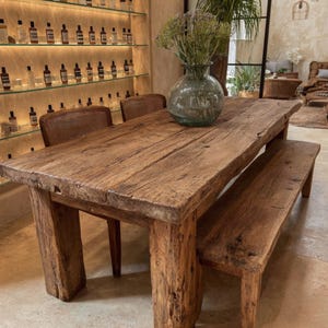 Peut inclure: Une table &agrave; manger rustique en bois avec un banc assorti. La table est faite de planches &eacute;paisses et patin&eacute;es et de pieds carr&eacute;s robustes. Un vase en verre transparent avec des fleurs s&eacute;ch&eacute;es est pos&eacute; sur la table. L'ensemble est en bois r&eacute;cup&eacute;r&eacute;.
