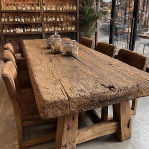 Peut inclure: Une grande table &agrave; manger en bois rustique avec une finition vieillie. La table est soutenue par des pieds &eacute;pais et robustes et entour&eacute;e de chaises en cuir marron. Des bocaux en verre et des bougies sont pos&eacute;s sur la table. En arri&egrave;re-plan, des &eacute;tag&egrave;res remplies de bouteilles.