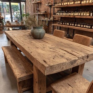 Peut inclure: Une grande table &agrave; manger en bois rustique avec un banc et des chaises assortis. La table est en bois r&eacute;cup&eacute;r&eacute;, mettant en valeur le grain et les n&oelig;uds naturels. Un vase d&eacute;coratif avec des plantes s&eacute;ch&eacute;es est pos&eacute; sur la table. La pi&egrave;ce a un mur de briques et des &eacute;tag&egrave;res avec des bouteilles.