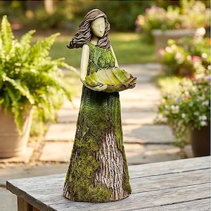 May include: A garden statue of a woman holding a large leaf-shaped bowl. The statue's dress is textured with green moss and bark-like details. The woman's hair is brown, and the bowl is a light green color. The statue is placed on a wooden surface.