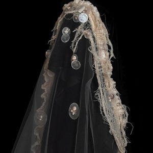 May include: A black veil featuring a beige frayed fabric trim along the edge. The veil is decorated with scattered translucent circular discs, small white pearls, and tiny seashells attached to the netting and the textured beige border.