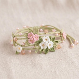 May include: A multi-strand bracelet made of sage green thread featuring crocheted pink and white flowers with green leaves. The design is accented with small pink pearls and translucent peach-colored beads interspersed along the thin cord strands.