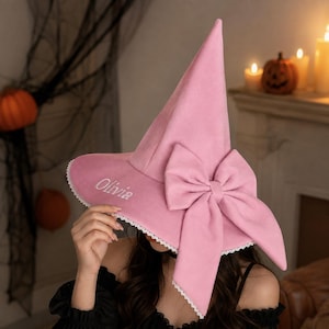 May include: A light pink pointed hat with a wide brim, featuring a large matching fabric bow on the side, white lace trim along the edge, and the name Olivia embroidered in white script on the front of the brim.