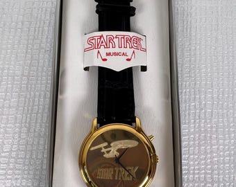 Reloj musical vintage de Star Trek en tono dorado con correa negra.