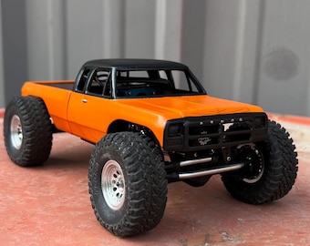May include: A bright orange remote-controlled pickup truck model featuring a black roof, a black front grille, and large, knobby off-road tires with silver rims. The vehicle has a lifted suspension system and a detailed chassis visible underneath.