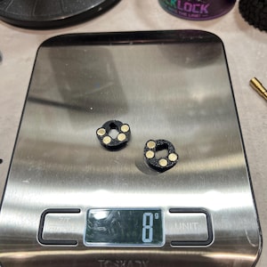 May include: Two small, black, circular components with three embedded gold-colored circular accents on each, resting on a digital scale that displays a weight of 8 grams. The components feature a central hole and a textured, matte black finish.
