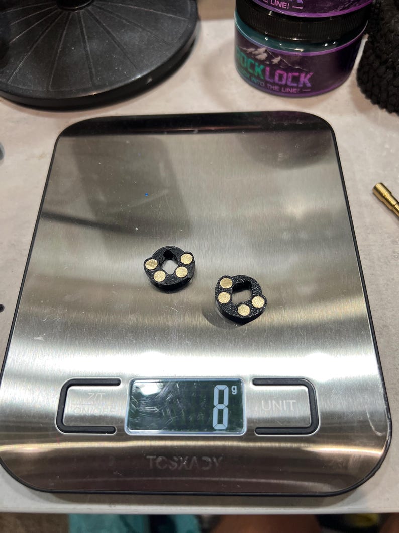 May include: Two small, black, circular components with three embedded gold-colored circular accents on each, resting on a digital scale that displays a weight of 8 grams. The components feature a central hole and a textured, matte black finish.