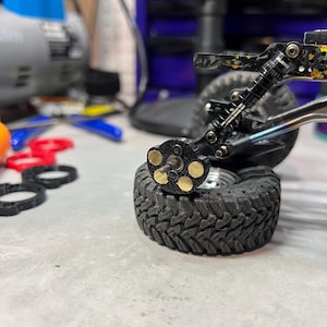 May include: A remote control vehicle wheel assembly featuring a black tire with a deep tread pattern, a metallic axle, and a black hub component with five circular brass weights attached. The suspension system includes a black coil-over shock absorber.