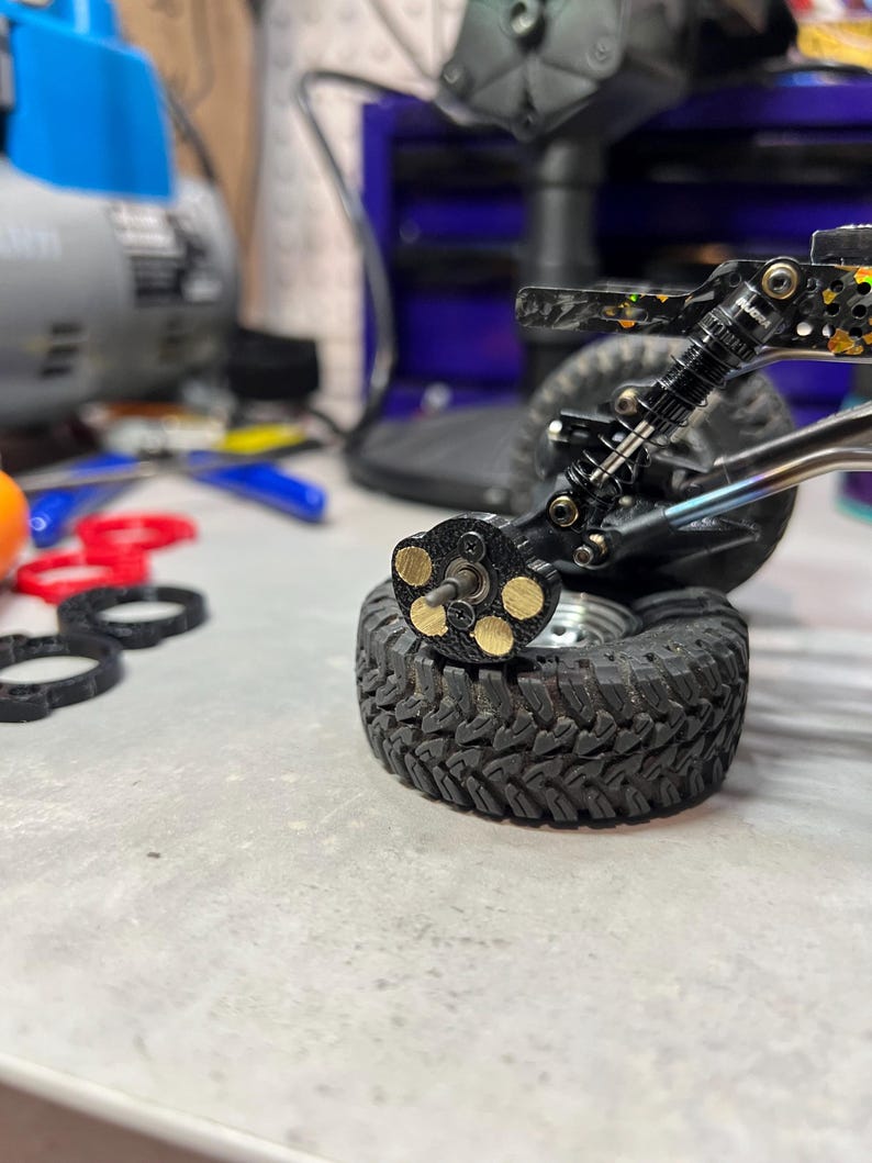 May include: A remote control vehicle wheel assembly featuring a black tire with a deep tread pattern, a metallic axle, and a black hub component with five circular brass weights attached. The suspension system includes a black coil-over shock absorber.