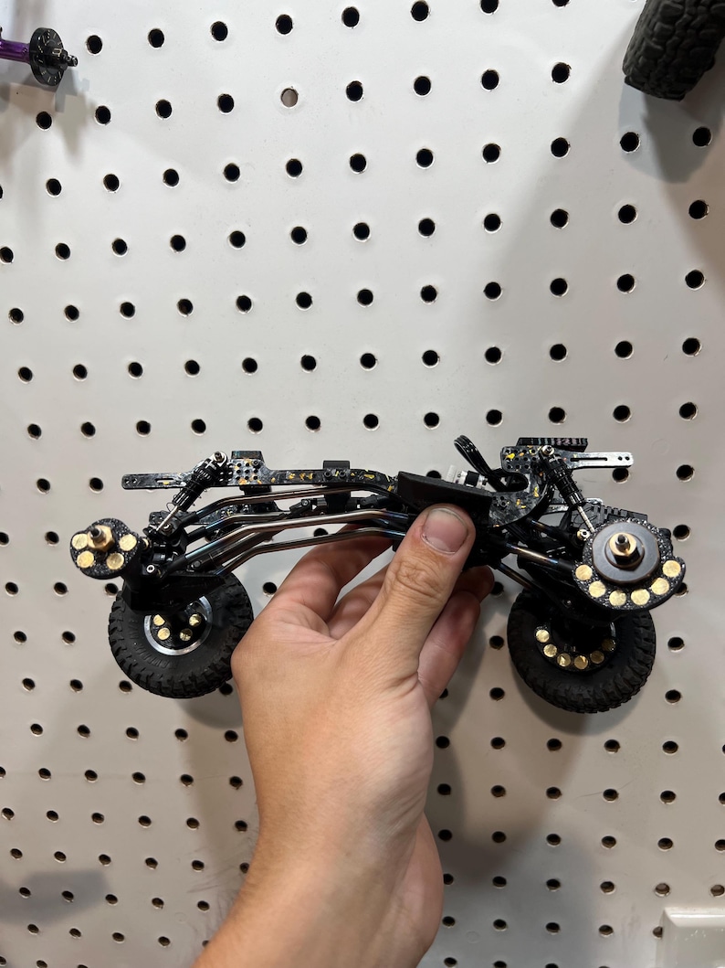 May include: A black and gold speckled remote control car chassis featuring a complex suspension system, four black rubber tires with gold-accented wheel hubs, and a metallic frame structure designed for high-performance off-road model racing.