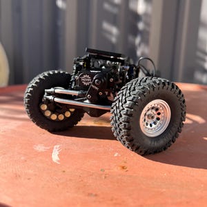 May include: A remote-controlled crawler chassis featuring large black off-road tires with deep treads, silver beadlock wheels, and a black central motor housing marked with the NSDRC logo. The vehicle includes exposed metal suspension links.