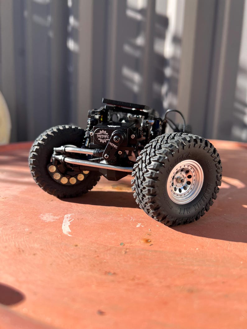 May include: A remote-controlled crawler chassis featuring large black off-road tires with deep treads, silver beadlock wheels, and a black central motor housing marked with the NSDRC logo. The vehicle includes exposed metal suspension links.