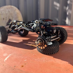 May include: A black radio-controlled crawler chassis featuring a detailed suspension system, exposed metal linkages, and knobby rubber tires. The frame includes a motor, shock absorbers, and brass-colored wheel hubs with visible circular cutouts.