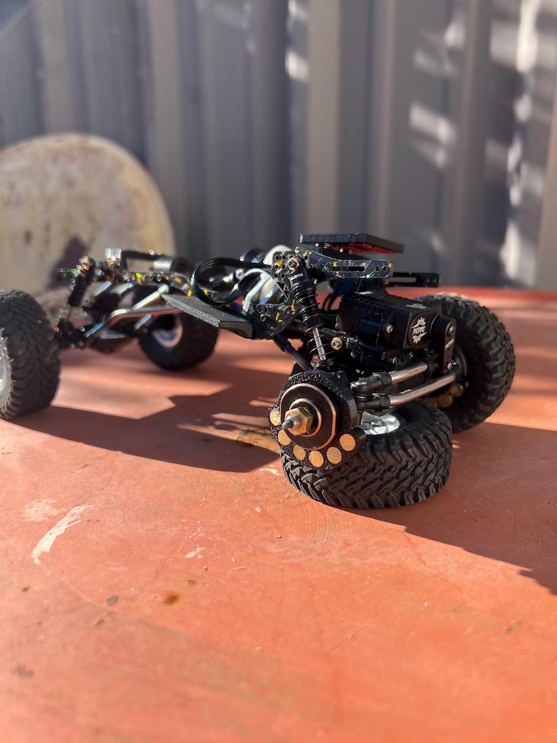 May include: A black radio-controlled crawler chassis featuring a detailed suspension system, exposed metal linkages, and knobby rubber tires. The frame includes a motor, shock absorbers, and brass-colored wheel hubs with visible circular cutouts.