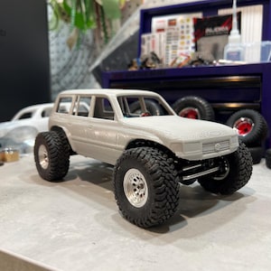 Può includere: Un modellino in scala di un SUV stampato in 3D di colore grigio chiaro con un design fuoristrada robusto. Il veicolo presenta grandi pneumatici in gomma nera con battistrada profondo montati su cerchi argentati, una griglia anteriore dettagliata.
