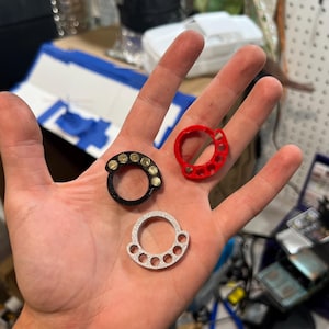 May include: Three circular rings with four small finger holes along the top edge. One ring is black with gold-colored circular accents, one is solid red, and one is silver with a textured surface. Each ring features a large central opening.