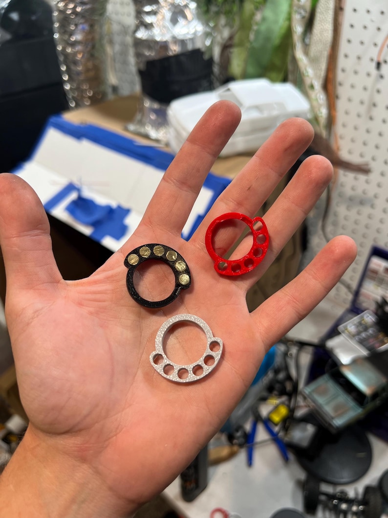 May include: Three circular rings with four small finger holes along the top edge. One ring is black with gold-colored circular accents, one is solid red, and one is silver with a textured surface. Each ring features a large central opening.