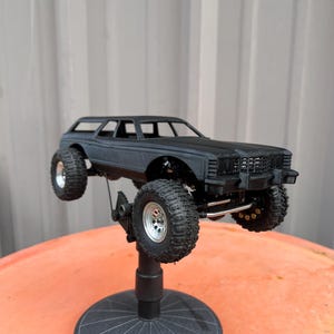 Puede incluir: Un modelo a escala de coche familiar en color negro mate con neumáticos todoterreno grandes y texturizados y llantas plateadas. El vehículo está montado sobre un soporte de exhibición circular negro, mostrando su chasis elevado, parrilla y ventanas.