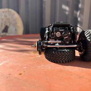 May include: A black remote-controlled crawler vehicle chassis featuring a prominent black servo motor with the white text NSDRC printed on the casing. The vehicle includes black off-road tires with deep treads, silver metal suspension links, and a black frame.