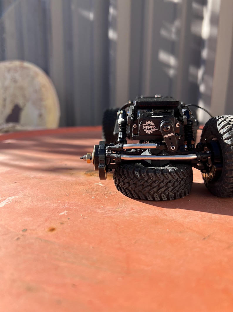 May include: A black remote-controlled crawler vehicle chassis featuring a prominent black servo motor with the white text NSDRC printed on the casing. The vehicle includes black off-road tires with deep treads, silver metal suspension links, and a black frame.