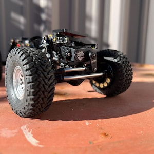 May include: A black remote-controlled crawler vehicle featuring large, deep-treaded tires with silver rims, a visible suspension system, and a central black motor housing marked with the text NSPRC and a gear icon, set on a flat surface.