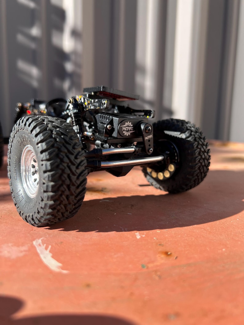 May include: A black remote-controlled crawler vehicle featuring large, deep-treaded tires with silver rims, a visible suspension system, and a central black motor housing marked with the text NSPRC and a gear icon, set on a flat surface.