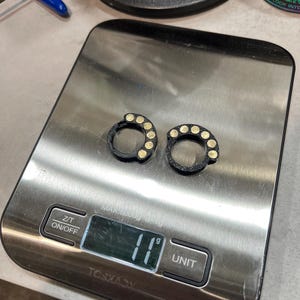 May include: Two black circular rings with five small gold-colored circular inlays on each, resting on a digital kitchen scale that displays a weight of 11 grams. The scale features a stainless steel surface with Z/T and ON/OFF buttons.