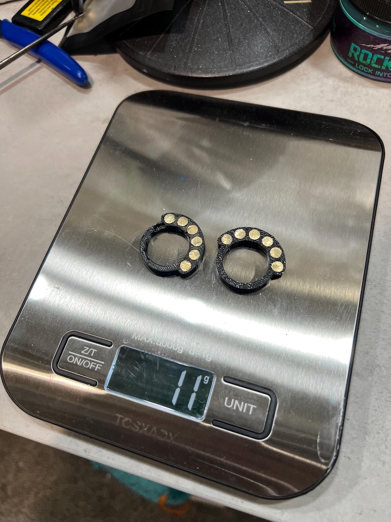 May include: Two black circular rings with five small gold-colored circular inlays on each, resting on a digital kitchen scale that displays a weight of 11 grams. The scale features a stainless steel surface with Z/T and ON/OFF buttons.