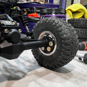 May include: A black remote-controlled crawler vehicle featuring a prominent black rubber tire with deep treads mounted on a silver metal rim. The wheel hub is secured with three visible brass-colored circular fasteners. The suspension system includes a black coil-over shock absorber attached to the axle assembly.