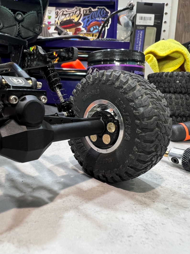 May include: A black remote-controlled crawler vehicle featuring a prominent black rubber tire with deep treads mounted on a silver metal rim. The wheel hub is secured with three visible brass-colored circular fasteners. The suspension system includes a black coil-over shock absorber attached to the axle assembly.