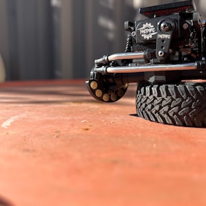 May include: A black remote-controlled vehicle axle assembly featuring a servo motor with the white text NSDRC printed on it. The component includes silver metal steering linkages, a black tire with a deep tread pattern, and a circular wheel hub.