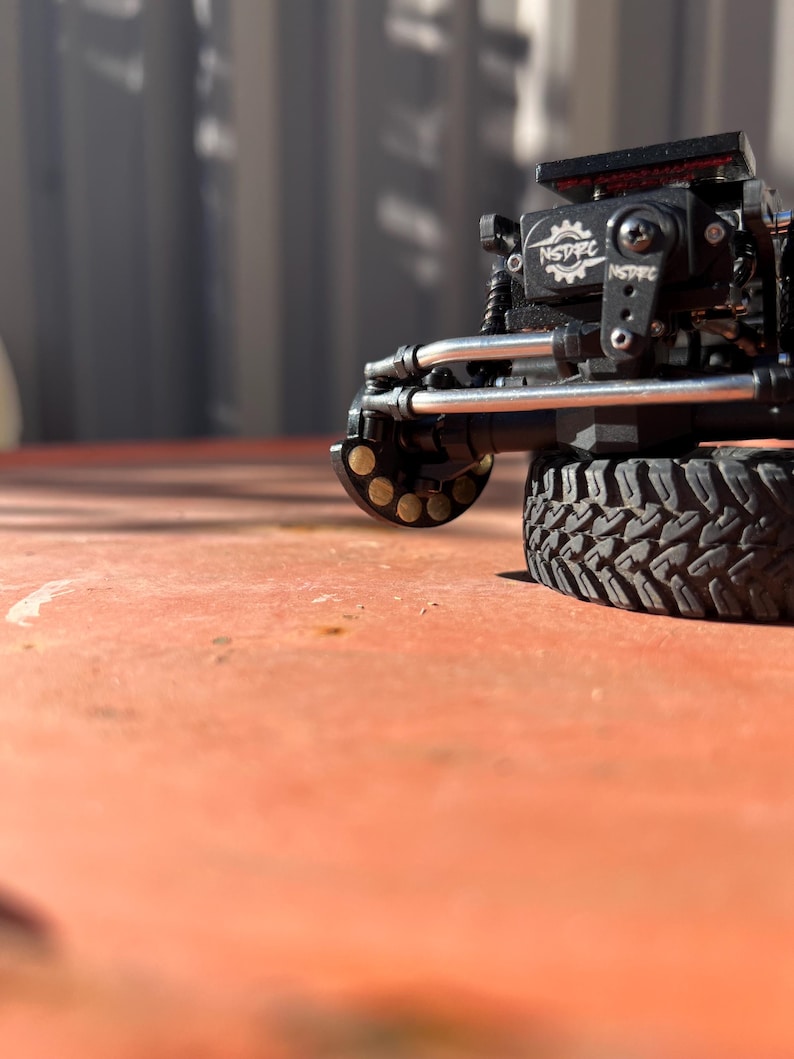 May include: A black remote-controlled vehicle axle assembly featuring a servo motor with the white text NSDRC printed on it. The component includes silver metal steering linkages, a black tire with a deep tread pattern, and a circular wheel hub.