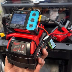 May include: A SkyRC B6neo 2 battery charger with a blue control panel attached to a red and black Milwaukee M18 RedLithium XC4.0 battery pack. The charger features a digital display screen, navigation buttons, and a connected small black battery.