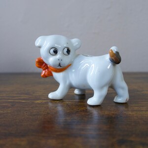 May include: A white ceramic dog figurine featuring large black eyes, a small nose, and a bright orange bow tied around its neck. The dog has a brown-tipped tail and is standing in a profile position, showcasing a smooth, glossy finish throughout.