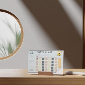 Può includere: Un grafico per il vasino con un camion e un escavatore, esposto su un supporto di legno. Il grafico ha caselle per tenere traccia dei progressi e le parole "Potty Chart" in alto. Una penna bianca poggia sulla superficie di legno.