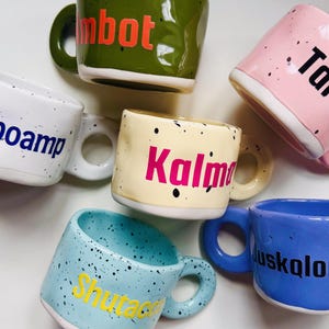 May include: A collection of five ceramic mugs in olive green, light pink, white, cream, and light blue, each featuring black speckles and a unique word printed in a bold font: mbot, Tar, poamp, Kalm, and Shutac, with a blue mug labeled juskolo.