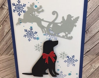 May include: A white card with a blue border features a black silhouette of a dog wearing a red bow. The background is decorated with blue and white snowflakes and a silver silhouette of Santa Claus in a sleigh pulled by reindeer.