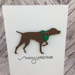 Peut inclure: Carte de Noël blanche avec une silhouette de chien marron portant un collier de houx vert et rouge. Les mots "merry CHRISTMAS" sont écrits en script noir sous le chien.