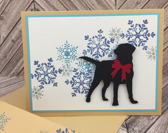 May include: A black silhouette of a dog wearing a red bow tie, surrounded by blue and gray snowflakes on a white background. The card has a light brown border and a matching envelope with a snowflake pattern.
