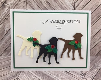 May include: A Christmas card with a white background and a green border. The card features three Labrador Retriever silhouettes in cream, black, and brown, each adorned with holly and a red gem. The words "merry CHRISTMAS" are written in black script.