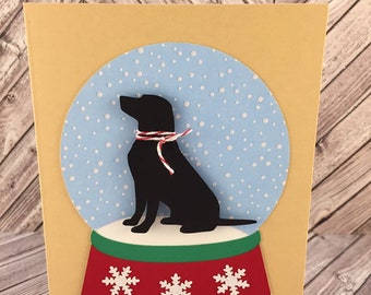 May include: A handmade greeting card featuring a black Labrador silhouette inside a snow globe. The snow globe has a light blue background with white dots, and a red and green base with white snowflakes. The card is a light yellow color.