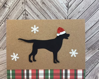 May include: A brown card featuring a black silhouette of a dog wearing a red glitter Santa hat. The card is decorated with white snowflake cutouts and a plaid patterned strip in red, green, and white.