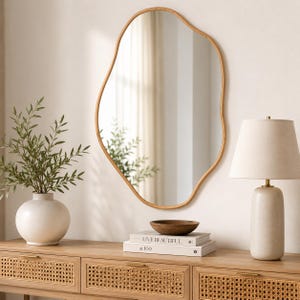 May include: An irregular, organic-shaped wall mirror with a light wood frame. The mirror reflects a room interior. Below it sits a stack of two books titled "LIVE BEAUTIFUL" and "at 100", topped with a small, round, dark brown wooden bowl.