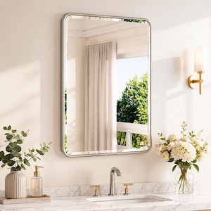 May include: A rectangular wall mirror with rounded corners and a silver frame. The mirror reflects a room with a white curtain and green trees outside. It is mounted above a white marble countertop with a silver faucet and two gold handles.