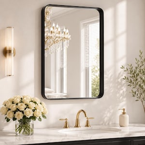 May include: A rectangular wall mirror with a thin black frame featuring rounded corners. The mirror reflects a crystal chandelier and a window with blinds. It is mounted above a white marble vanity countertop with a gold faucet and sink.