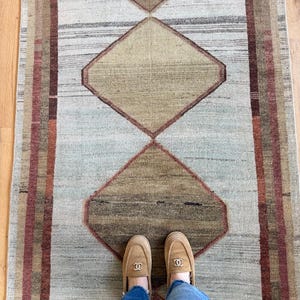 May include: A rectangular rug with a geometric diamond pattern in shades of beige, brown, and muted green. The rug has a striped border in red and brown. The rug is made of woven material.