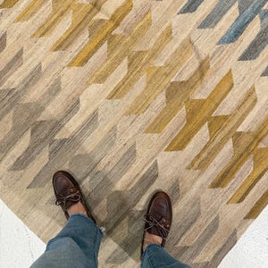 Puede incluir: Una alfombra estampada en tonos beige, dorado y azul. La alfombra presenta un diseño repetitivo de formas angulares. Zapatos náuticos de cuero marrón y vaqueros azules son visibles en primer plano.