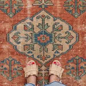 Peut inclure: Un tapis rectangulaire orné d'un médaillon géométrique central dans des tons orange rouille, bleu sourd et vert sauge. Le tapis présente une bordure décorative avec des motifs floraux répétitifs et une texture vieillie et usée.